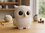 White knitted textured 3D printed owl figure/ key tag on a wooden surface with yarn and knitting needles in the background.